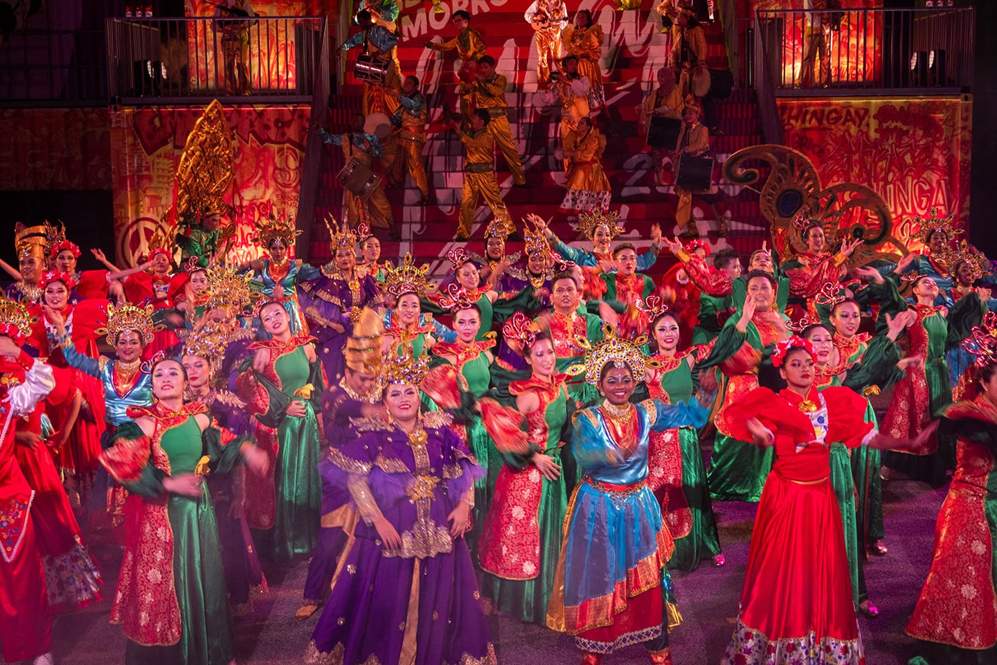 Large group onstage in colorful costumes and ornate headdresses, drummers in background.